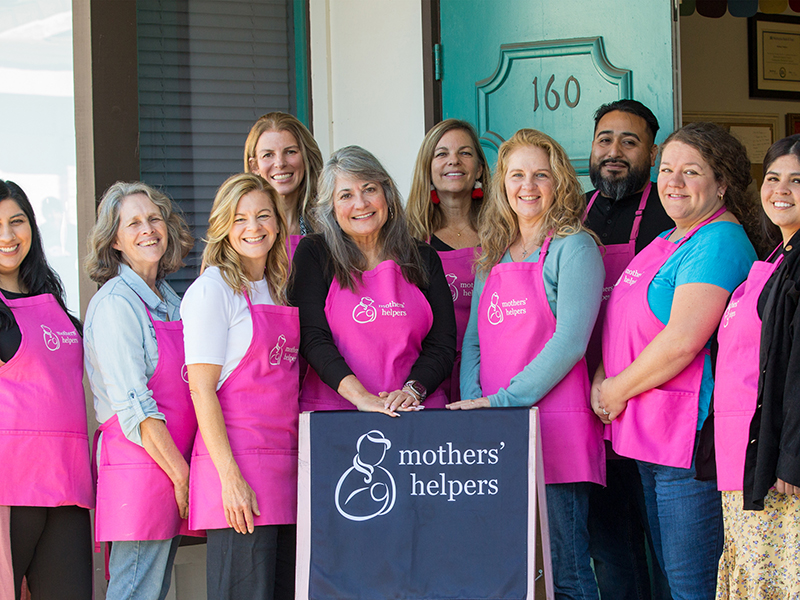 Volunteers with pink aprons behind mothers' helpers sign
